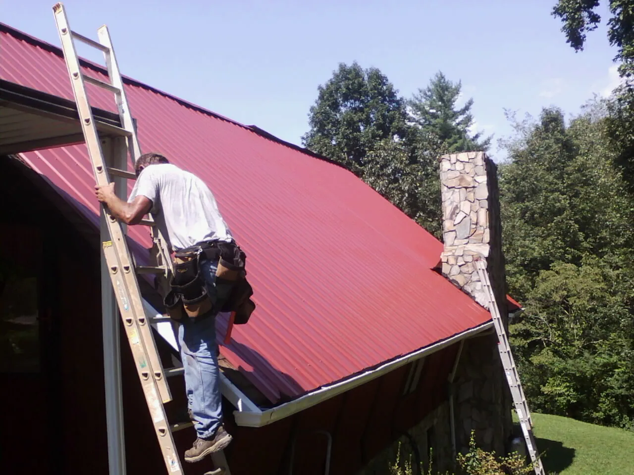 Roofer on a ladder beside a red metal roof and stone chimney — Soffit & Fascia Repair by Roofing Brothers in Adger